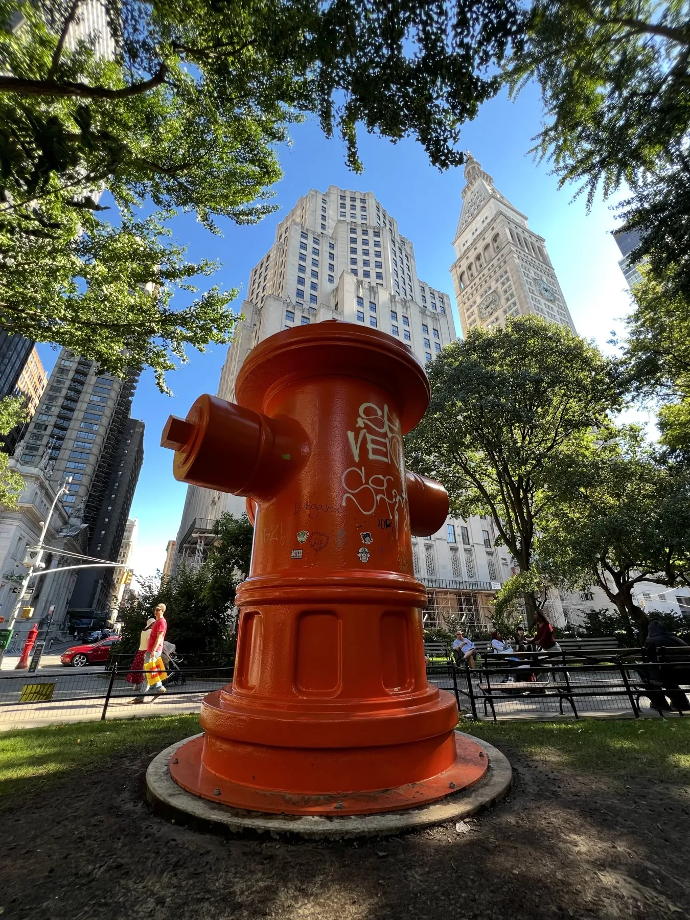 A bright orange hydrant-shaped sculpture framed by leafy trees and New York buildings