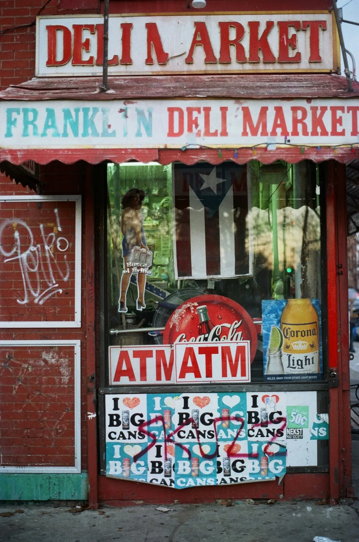 Weathered red deli storefront with layered signs and a Puerto Rican flag in the window