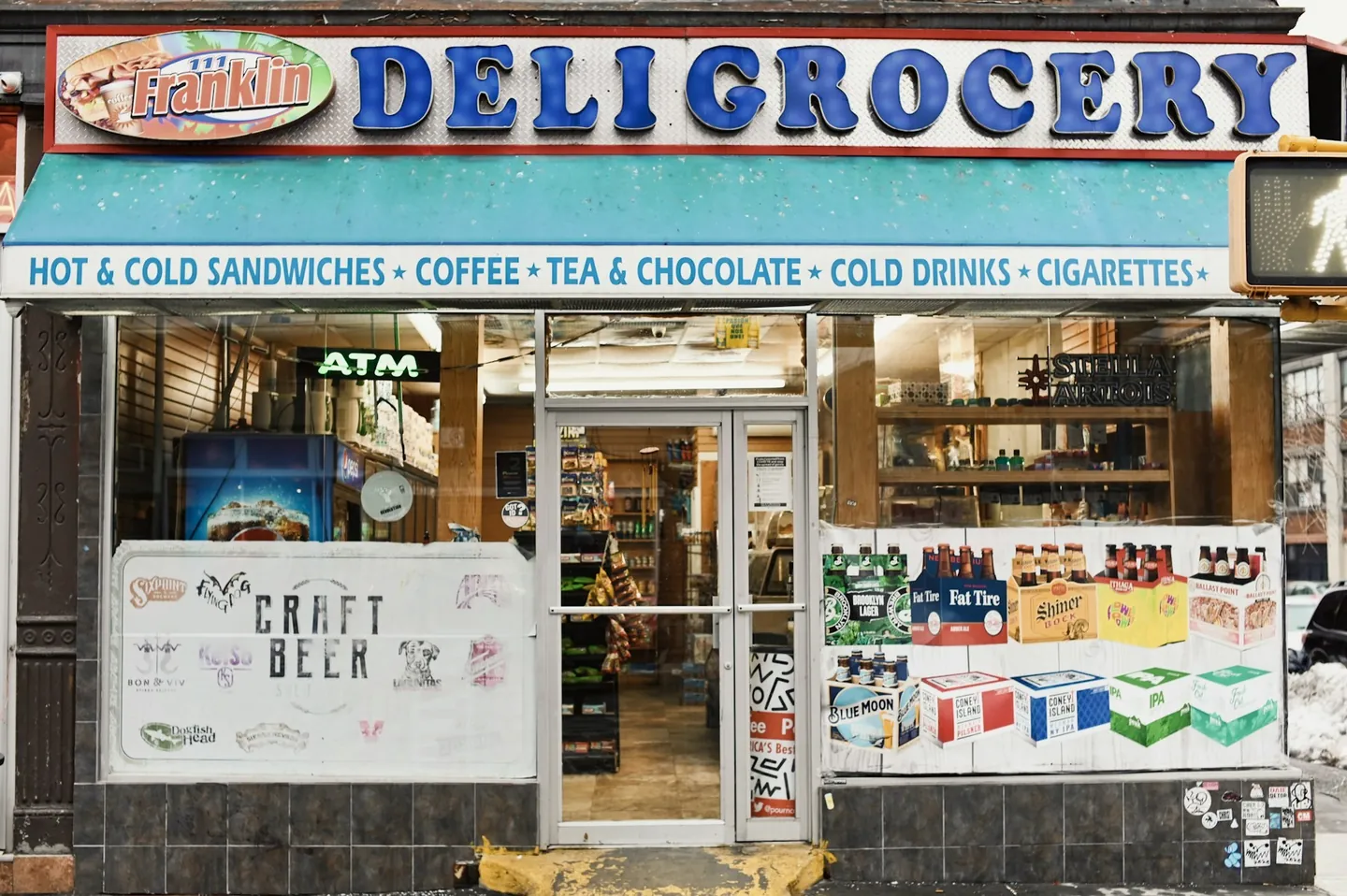 Bright Franklin Deli Grocery storefront with a teal awning, bold blue lettering, layered window signs and a yellow doorstep